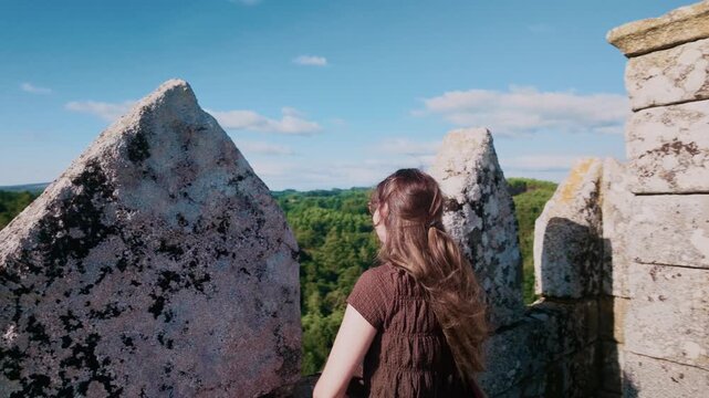 Young woman with glasses admiring the forest from the battlements of Pambre Castle in Galicia, Spain. She turns around, smiles, and her hair blows in the wind on a bright, sunny day