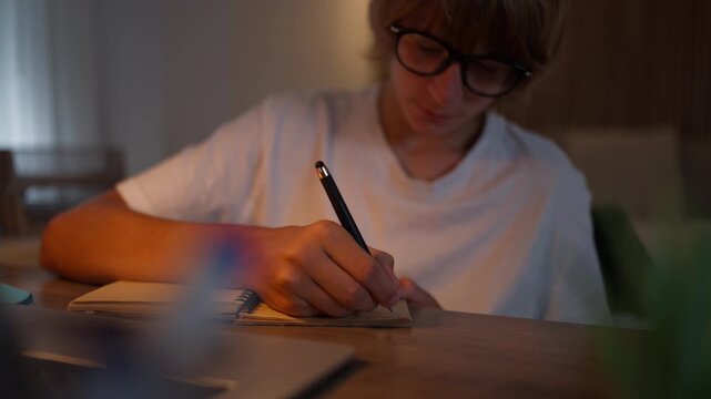 Focused teenager wearing glasses and taking handwritten notes in a notebook during a study session. Note-taking, learning, education, memory, concentration and academic skills concept.