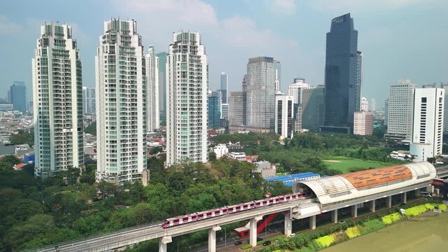 Jakarta, Indonesia - 17 July 2026: Aerial view of a modern train moving along an elevated track in front of the Jakarta skyline with tall skyscrapers and lush green trees.
