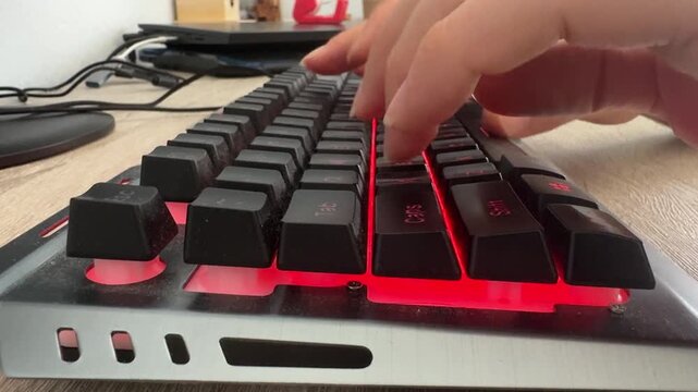 Side angle close-up of human hands efficiently operating a sleek black electronic keyboard featuring vibrant red led lights resting on a wooden surface highlighting daily modern computer interactions.