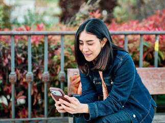 A young woman sits on a wooden park bench, smiling as she scrolls and texts on her smartphone. Dressed in a denim jacket with a leather backpack beside her, she looks relaxed and engaged in a candid u © Lumos sp