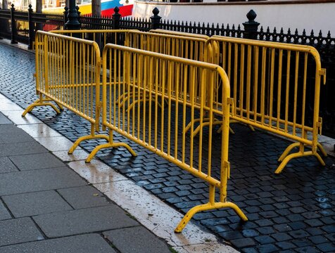 Yellow metal crowd control barriers stand on wet cobblestone sidewalk near black iron fence. Portable safety barricades line city street pavement for security event management. Weathered construction