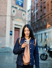 Fototapeta premium A young woman wearing a denim jacket checks her smartphone while standing on a city street. Young woman smiling while holding smartphone and looking up in an urban street