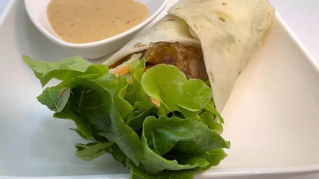 Overhead food video of tortilla wrap filled with fresh lettuce and savory filling, served with creamy dipping sauce on a white plate and clean background
