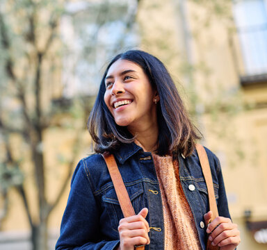 A young woman walks through a sunlit city neighborhood, smiling and looking upward with a confident, optimistic expression. She wears a denim jacket, cozy sweater and carries a leather backpack, conve