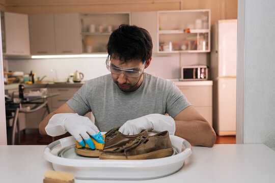 Focused man wearing safety glasses and gloves cleaning dirty brown footwear with sponge. Domestic chore and shoe care concept in modern kitchen background. Household maintenance.