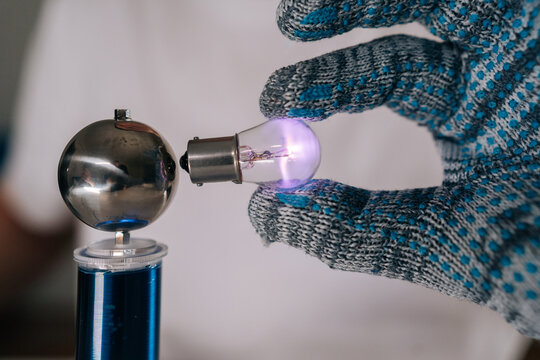 Close-up view of scientist hand in protective glove holding glowing light bulb near high frequency coil, demonstrating wireless electricity transmission and energy innovation in physics experiment.