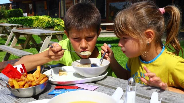 Two children eating soup together from shared bowl at outdoor table