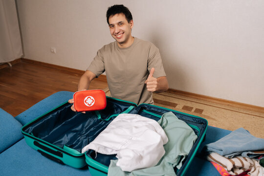 Portrait of happy traveler preparing for trip, placing travel medicine bag into suitcase filled with clothes, ensuring health and wellness while away from home, giving thumbs up looking at camera.