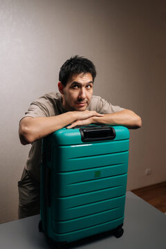 Pensive man leaning on a turquoise luggage, looking away and imagining future vacation getaway, feeling anticipation for departure and long-awaited travel adventures.