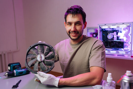Smiling technician showing dirty cooler fan before cleaning. Professional computer service and hardware maintenance concept. Workshop with tools and glowing PC in background.