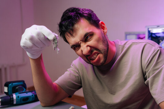 Intense portrait of frustrated man with angry expression cleaning dusty electronics. Person in workshop with neon light showing rage and disgust from dirty component.