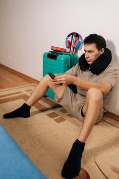 Young male traveler sitting on the floor with luggage, wearing a neck pillow and shorts, using a smartphone while waiting, conveying concepts of travel, anticipation, and delay.