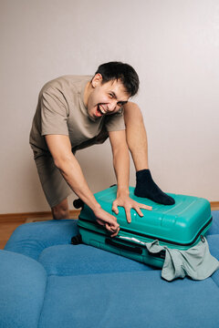 Vertical portrait of fury traveler male struggling to zip overpacked green suitcase for a trip, frowning and making a funny face, experiencing frustration and packing difficulty for vacation travels.