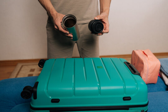 Cropped shot of traveler male holding reusable thermos and its lid above a turquoise suitcase, meticulously packing travel necessities while considering hydration for upcoming adventures.