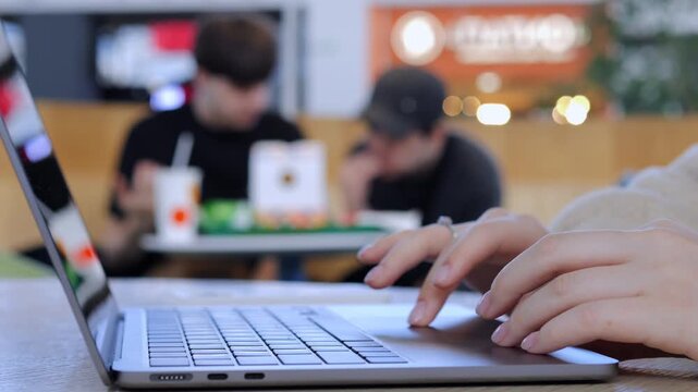 Close-up of hands typing on a laptop in a cafe. Background shows blurred people relaxing. Scene conveys focus, productivity, and casual work. Suitable for remote work, freelancing