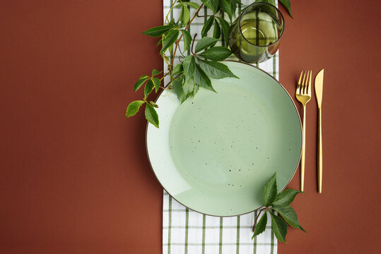 A thoughtfully arranged table setting featuring a light green speckled plate and golden cutlery next to a glass, adorned with fresh green leaves