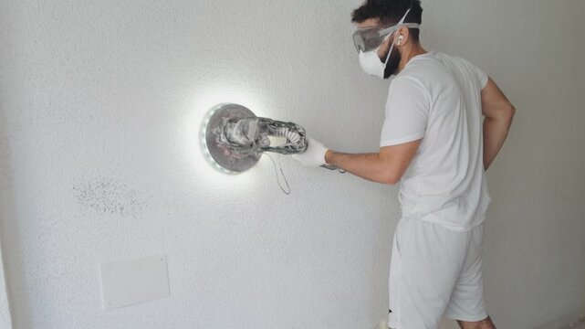 Male worker wearing protective safety mask and goggles using an electric orbital wall sander with bright LED light to smooth plaster and remove texture during home renovation and DIY construction