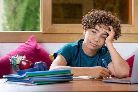 Teenage student sitting at a desk with a notebook and pen, looking tired, bored and unmotivated while studying at home. Concept of education, homework, academic stress, procrastination, lack of concen