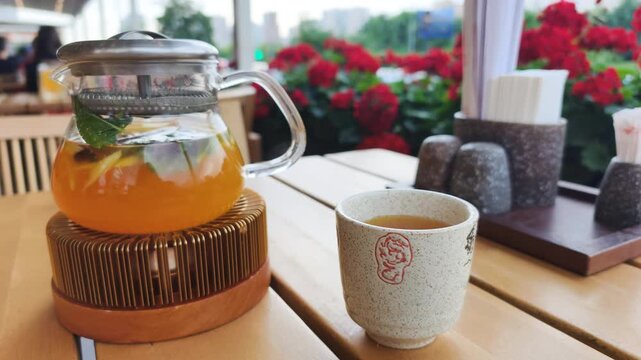 Hot buckthorn tea in glass teapot sits on heater stand near ceramic cup. Steaming buckthorn tea with ginger and citrus slices creates cozy drink serving on wooden cafe table outdoors.