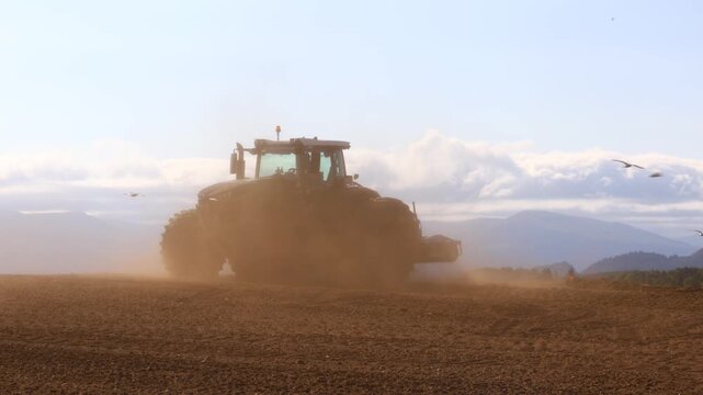 Large tractor plowing dry field in strong wind generating dense dust cloud across agricultural terrain