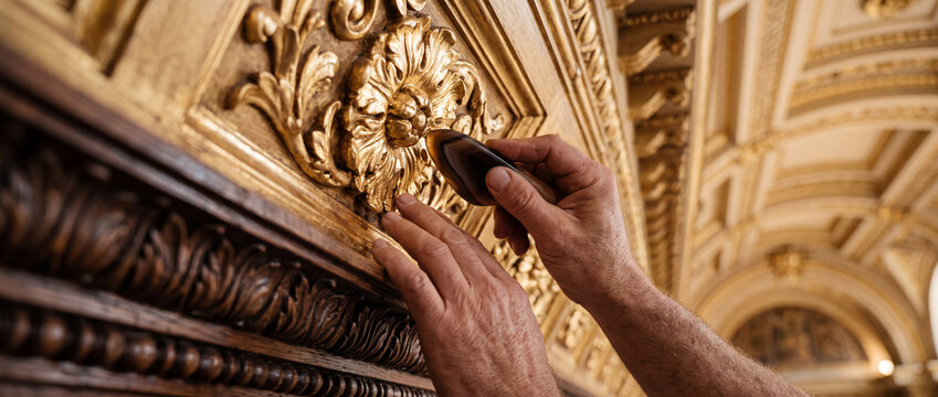 Artisan restoring gilded ornate ceiling detail with hands and tool in historic interior