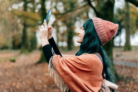 Young Woman wrapped in Shawl Poncho making photo by phone while enjoying autumn walk in forest. Capture the moment. Feeling harmony. Relaxing, personal fulfilment on nature. Local travel lifestyle.