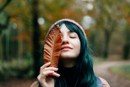 Young smiling woman with closed eyes covering her face with fallen leaf enjoying the moment of calm and freedom in autumn forest. Feeling harmony, reunion with nature. Relaxing, mental recovery.
