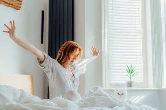 Stretching after the sun. Smiling relaxed middle aged woman stretching hands in bed after wake up in the morning in light sunny bedroom. Concept of a new day and joyful weekend.