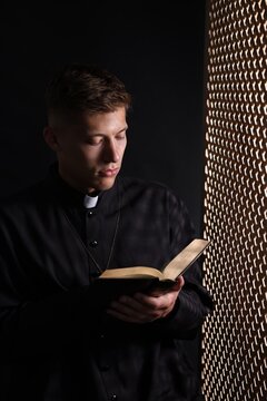 Priest with Holy Bible praying near partition in confessional booth