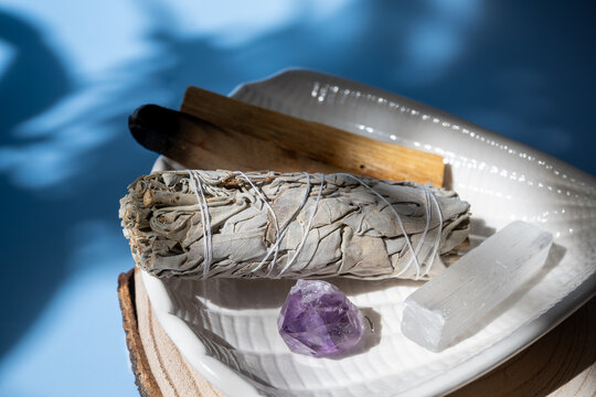 sage smudge stick and healing crystals on ceramic plate with shadow play on blue background