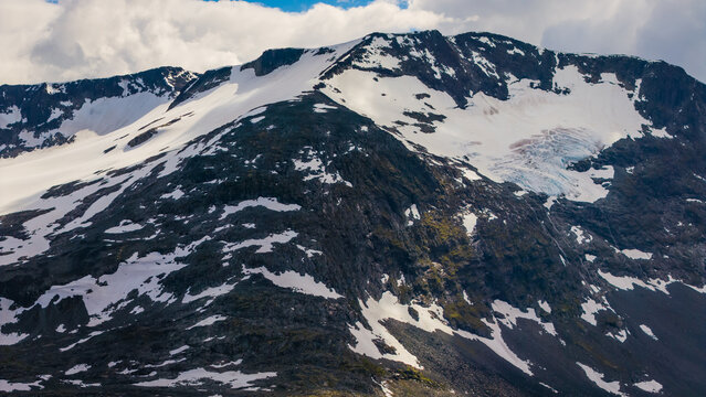 Aerial view of Sognefjellet mountain range with rugged rocky slopes and patches of snow under a cloudy sky in Lom Municipality, Innlandet, Norway.