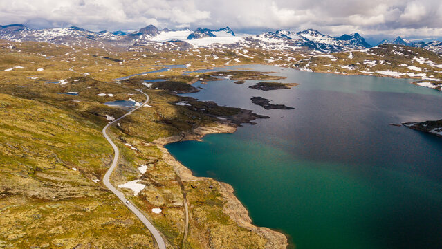 Aerial view of Sognefjellet mountain pass with a winding road, turquoise glacial lake, and snow-capped peaks under a cloudy sky in Lom Municipality, Innlandet, Norway.