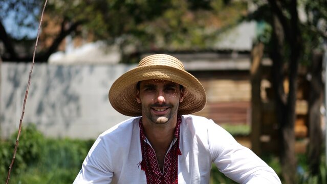 Portrait of ukrainian man in straw hat against background of summer garden. Young smiling guy wearing an embroidered shirt looking into camera outdoor. Concept of national identity and patriotism