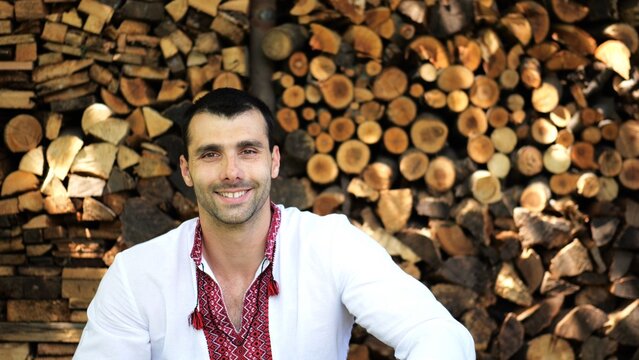 Portrait of ukrainian man against the background of woodshed. Young smiling guy wearing an embroidered shirt looking into camera outdoor. Concept of national identity and independence