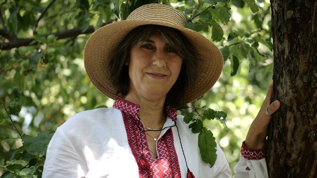 Portrait of ukrainian woman in straw hat against the background of summer garden. Adult smiling lady wearing an embroidered shirt looking into camera outdoor. National identity and patriotism concept