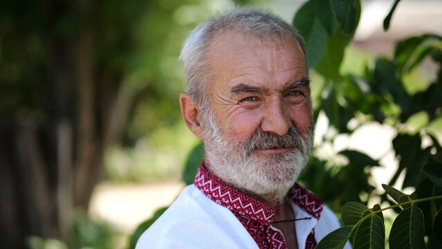 Portrait of old ukrainian man in vyshyvanka near green walnut tree of summer garden. Senior smiling male wearing an embroidered shirt looking into camera outdoor. National identity and patriotism