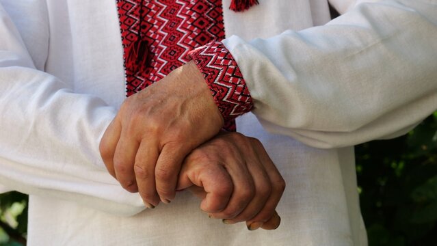 Hands of elderly ukrainian man in embroidered shirt rubbing together slowly outdoor. The movement is calm and thoughtful, symbolizing reflection, anticipation or simple presence in the moment