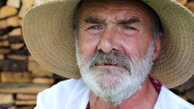 Portrait of senior ukrainian man in straw hat against the background of woodshed. Elderly serious male wearing an embroidered shirt looking into camera outdoor. Cultural heritage and ethnic roots