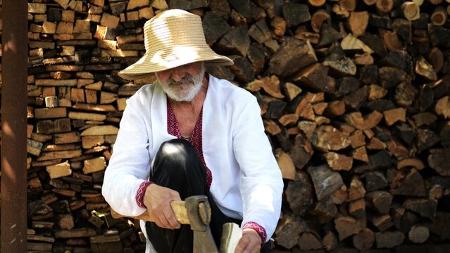 Older man wearing an embroidered shirt gently trims a piece of wood with axe against background of woodshed. Concept of craftsmanship, tradition and ukrainian heritage. Slow motion