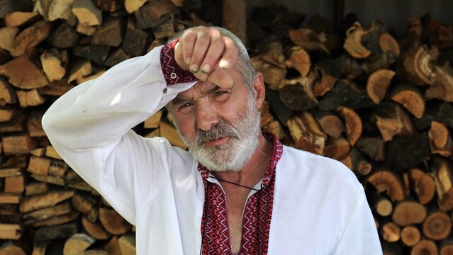 Senior ukrainian man in vyshyvanka standing after hard work near woodshed. Elder tired male resting after hard job near firewood. Concept of national identity and independence. Close up