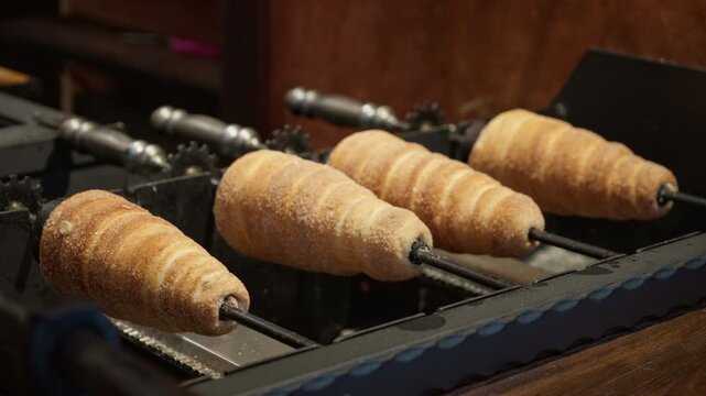 Traditional chimney cakes spin on spit rollers over an open grill. Sweet trdelnik pastry bakes into a delicious golden dessert. European street food vendor concept for festive holiday fairs.