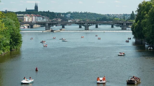 People ride paddle boats on Vltava River in Prague city. Tourists enjoy outdoor summer recreation on water. Historic bridge and city architecture expand in background for travel concept.