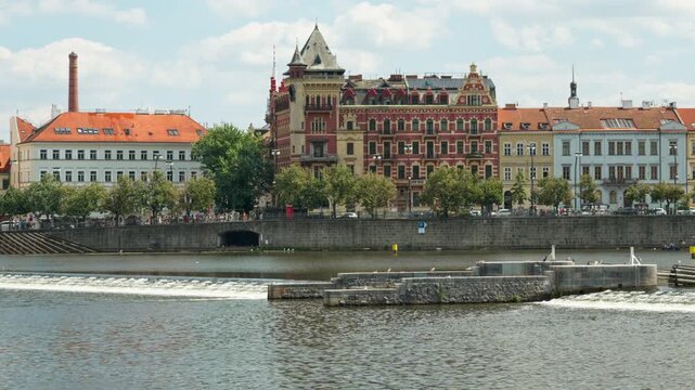 Vltava River weir flows near historic city embankment buildings in Prague. Water cascades over the urban dam structure. European travel destination concept for summer city break and tourism.