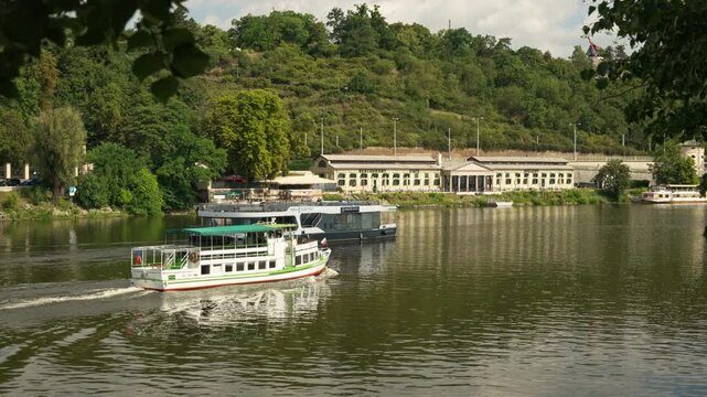 Tourist boats sail along Vltava River near green hill in Prague. Excursion passenger ships cruise on scenic water. Concept of European river travel, summer vacation, and sightseeing tour.
