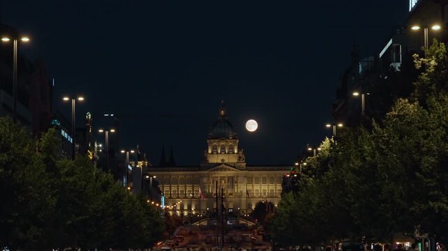 Full moon shines over illuminated National Museum on Wenceslas Square at night in Prague. Bright city lights glow along avenue with tourists. Concept of European urban night tourism.