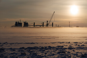 Industrial arctic landscape with cargo vessel and port crane hidden in thick sea smoke during frosty winter sunset. Northern Russia with copy space. © Konstantin