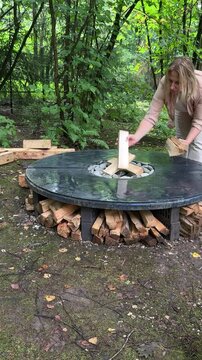 Woman adding split firewood to built-in storage underneath a handmade round concrete fire pit table with decorative stones in a woodland garden.