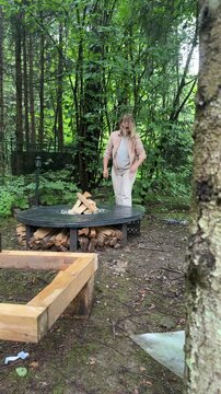 Woman adding split firewood to built-in storage underneath a handmade round concrete fire pit table with decorative stones in a woodland garden.