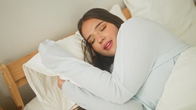 Woman hugging pillow with closed eyes on bed in a bedroom inside a building, wooden frame and white bedding visible; serenity.